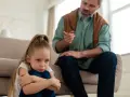 Smutna dziewczynka skulona na podłodze, podczas gdy ojciec mówi do niej z kanapy. To może być obraz pokazujący objawy daddy issues.