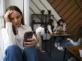 Dziewczyna z telefonem w dłoni, z ręką na czole, czuje się wyobcowana. W tle inne osoby, które nie zwracają na nią uwagi, odzwierciedlając jej neleczona fobia społeczna.