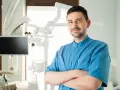 Uśmiechnięty dentysta w niebieskiej koszuli stoi ze skrzyżowanymi rękami w gabinecie stomatologicznym.