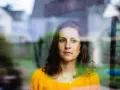 Kobieta w żółtym swetrze patrzy przez okno. Jej wyraz twarzy może być trudny do zinterpretowania, co rodzi pytania, jak rozpoznać osobę chorą psychicznie.