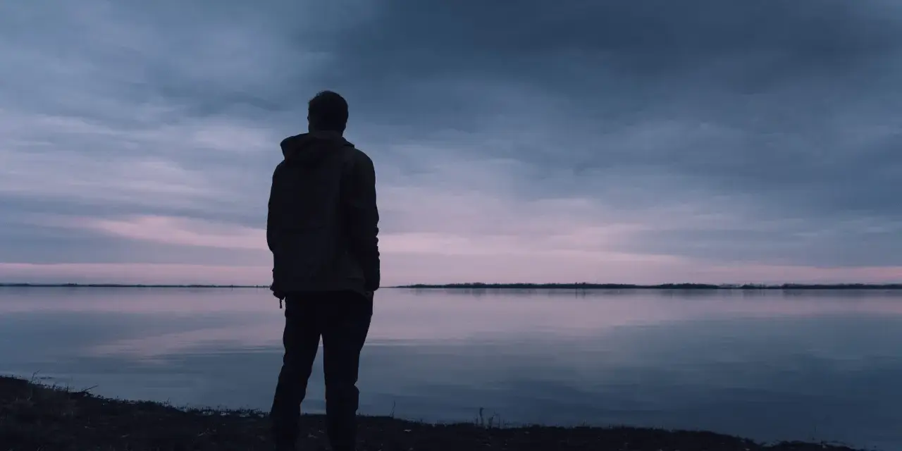 Samotny mężczyzna stoi nad jeziorem o zmierzchu, patrząc w dal. Czy to pesymista, kto to, zastanawia się nad mrocznym niebem i spokojną wodą?