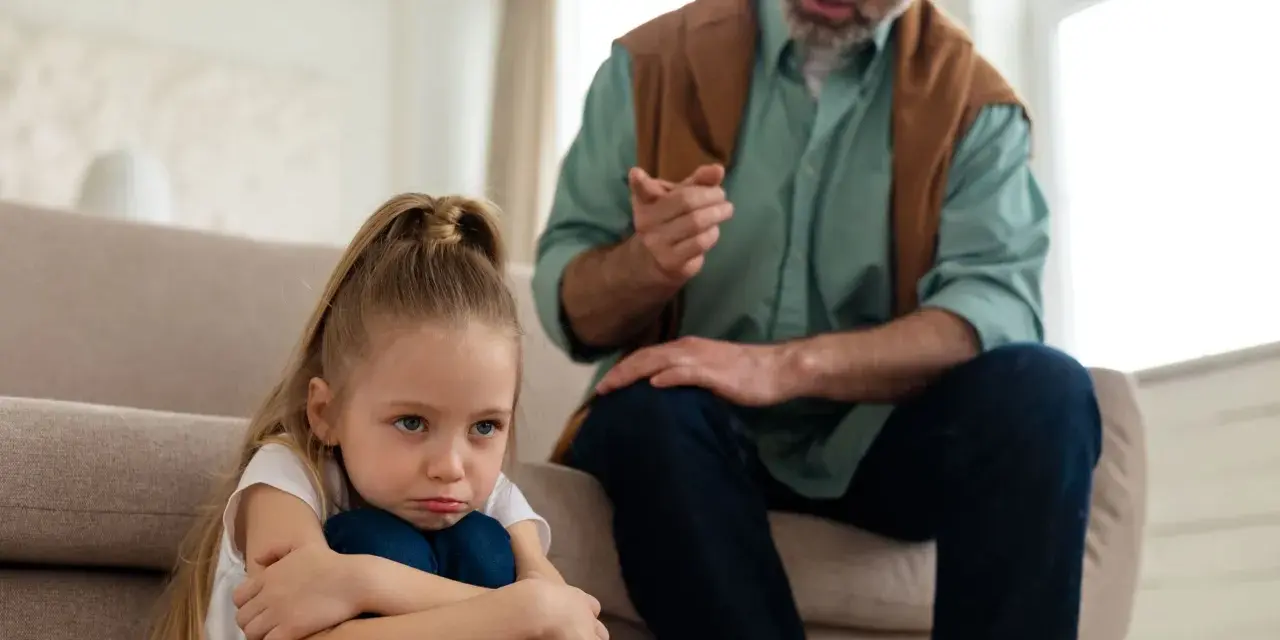Smutna dziewczynka skulona na podłodze, podczas gdy ojciec mówi do niej z kanapy. To może być obraz pokazujący objawy daddy issues.