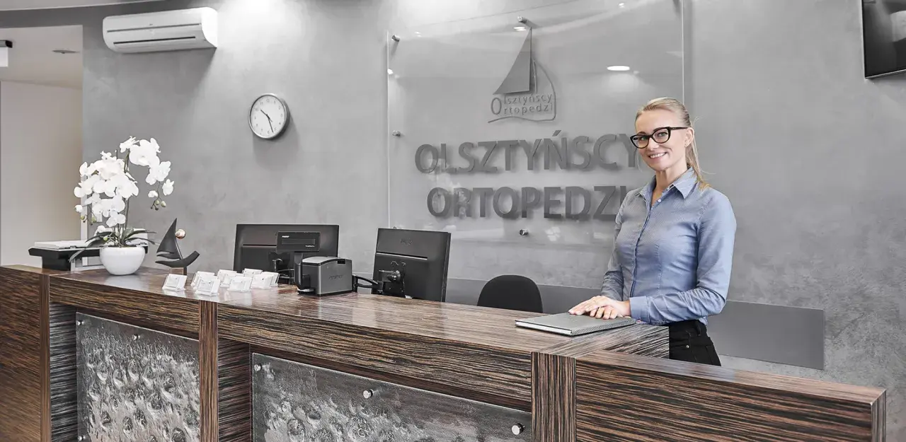 Uśmiechnięta recepcjonistka w okularach za ladą w gabinecie ortopedycznym.