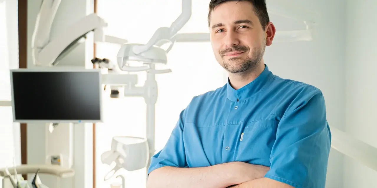 Uśmiechnięty dentysta w niebieskiej koszuli stoi ze skrzyżowanymi rękami w gabinecie stomatologicznym.