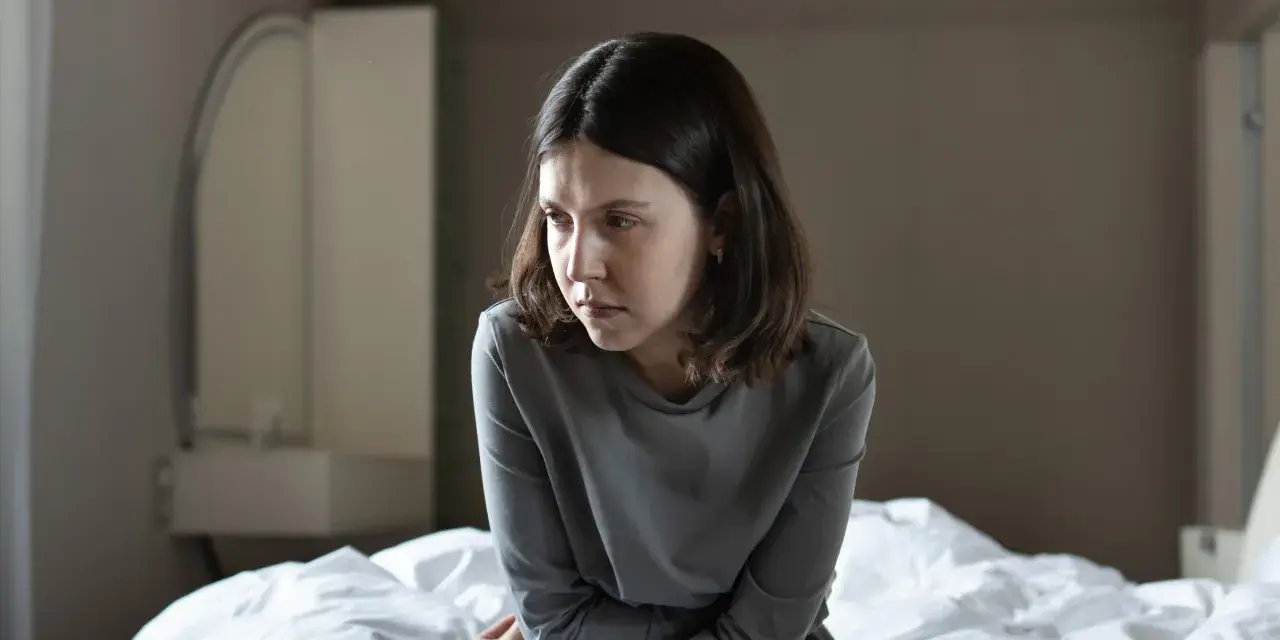Młoda kobieta siedzi na łóżku, zamyślona. Jej wyraz twarzy sugeruje walkę z depresją, ale w jej oczach widać iskierkę nadziei na lepsze jutro.