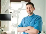 Uśmiechnięty dentysta w niebieskiej koszuli stoi ze skrzyżowanymi rękami w gabinecie stomatologicznym.