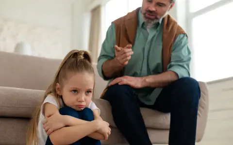 Smutna dziewczynka skulona na podłodze, podczas gdy ojciec mówi do niej z kanapy. To może być obraz pokazujący objawy daddy issues.