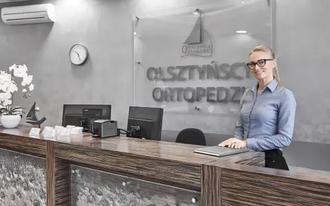 Uśmiechnięta recepcjonistka w okularach za ladą w gabinecie ortopedycznym.
