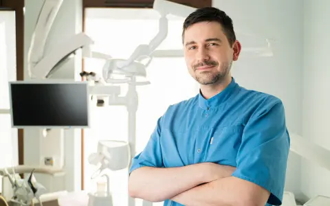 Uśmiechnięty dentysta w niebieskiej koszuli stoi ze skrzyżowanymi rękami w gabinecie stomatologicznym.