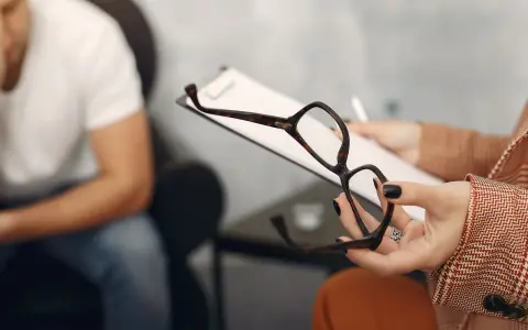 Psycholog a psychoterapeuta różnice: sesja terapeutyczna, gdzie terapeuta trzyma okulary i notatki, a pacjent siedzi w tle.