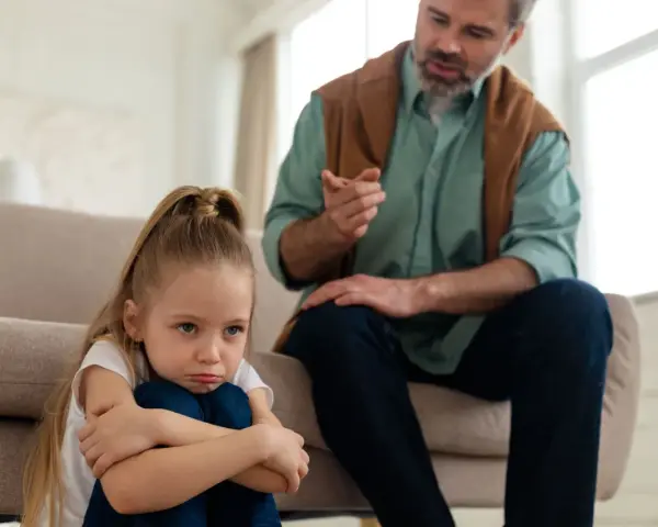 Smutna dziewczynka skulona na podłodze, podczas gdy ojciec mówi do niej z kanapy. To może być obraz pokazujący objawy daddy issues.