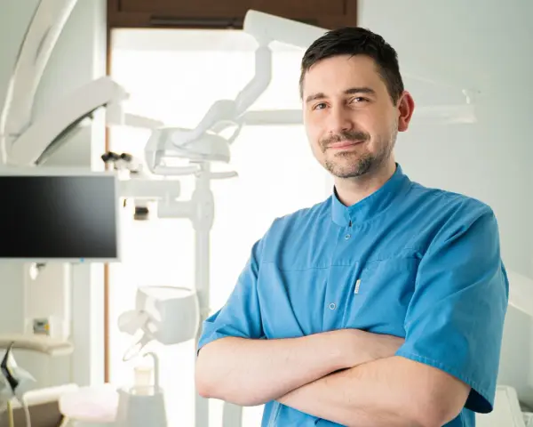 Uśmiechnięty dentysta w niebieskiej koszuli stoi ze skrzyżowanymi rękami w gabinecie stomatologicznym.