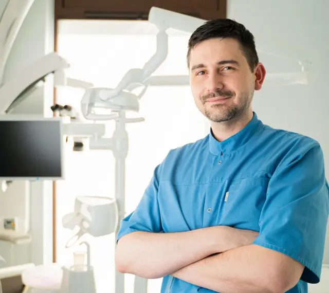 Uśmiechnięty dentysta w niebieskiej koszuli stoi ze skrzyżowanymi rękami w gabinecie stomatologicznym.