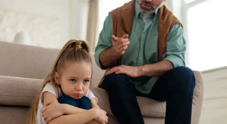 Smutna dziewczynka skulona na podłodze, podczas gdy ojciec mówi do niej z kanapy. To może być obraz pokazujący objawy daddy issues.