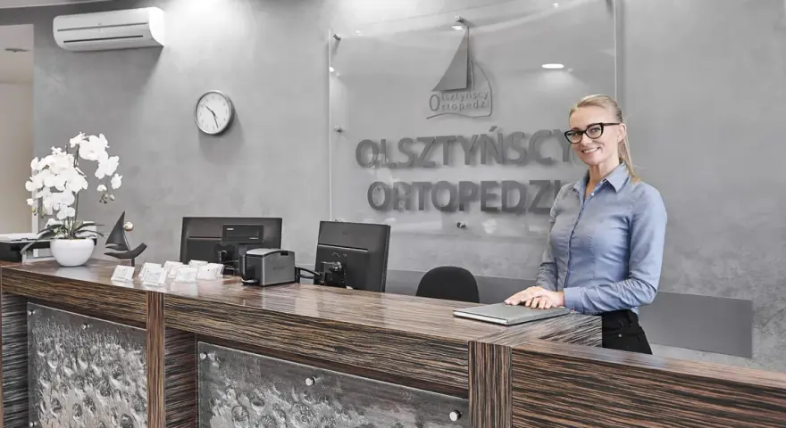 Uśmiechnięta recepcjonistka w okularach za ladą w gabinecie ortopedycznym.