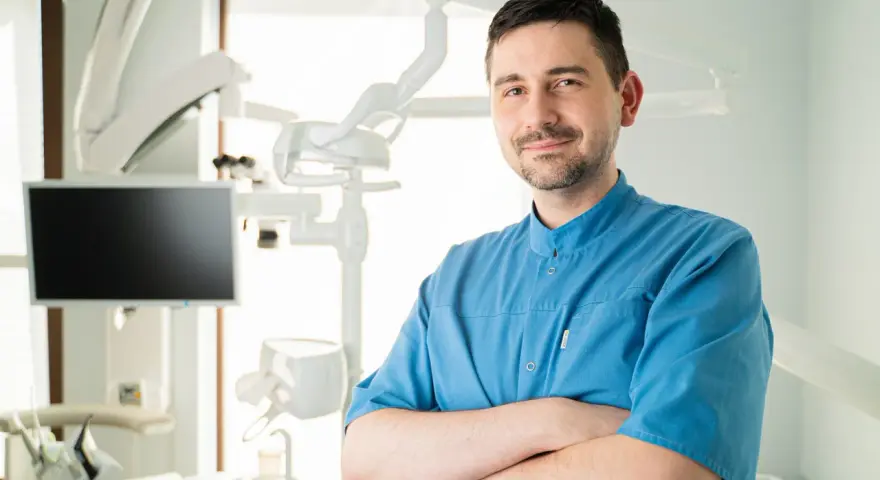 Uśmiechnięty dentysta w niebieskiej koszuli stoi ze skrzyżowanymi rękami w gabinecie stomatologicznym.