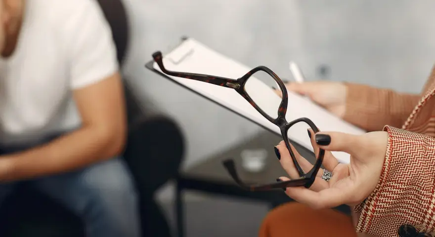 Psycholog a psychoterapeuta różnice: sesja terapeutyczna, gdzie terapeuta trzyma okulary i notatki, a pacjent siedzi w tle.