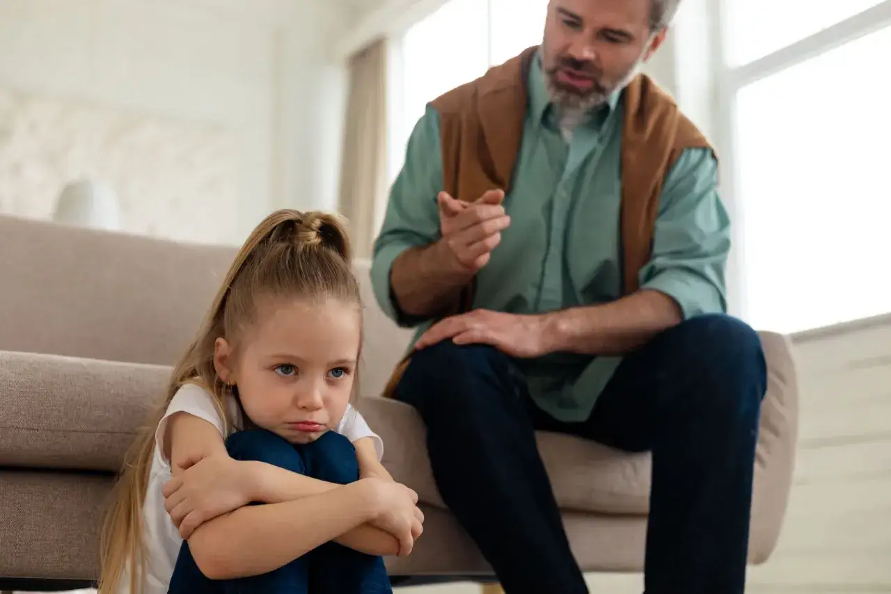 Smutna dziewczynka skulona na podłodze, podczas gdy ojciec mówi do niej z kanapy. To może być obraz pokazujący objawy daddy issues.