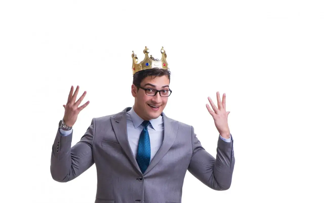 Mężczyzna w garniturze i koronie, z uniesionymi rękami, wygląda jak megaloman. Kto to?