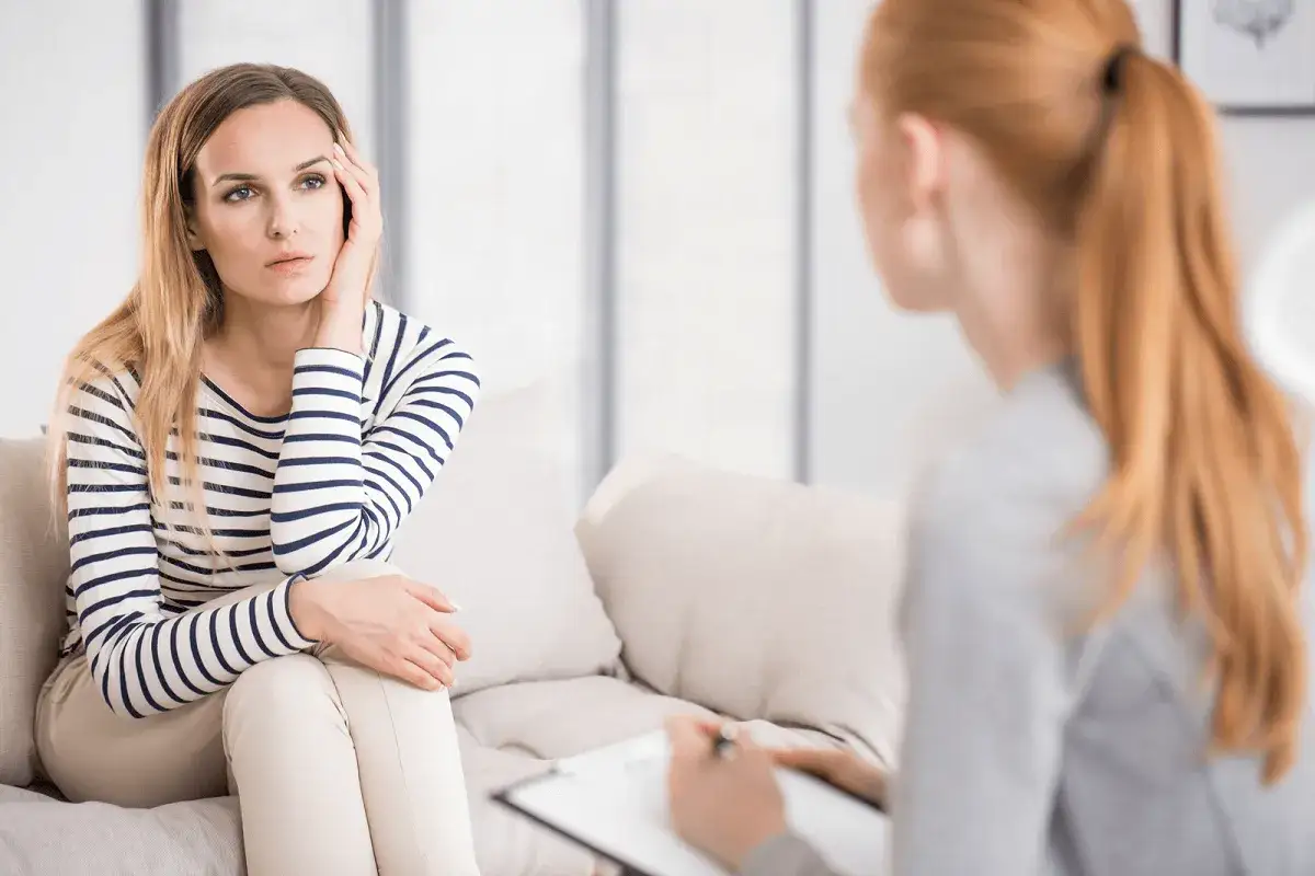 Kobieta w paski rozmawia z psychologiem, który notuje. Widać, co robi psycholog, gdy pacjentka opowiada o swoich problemach.