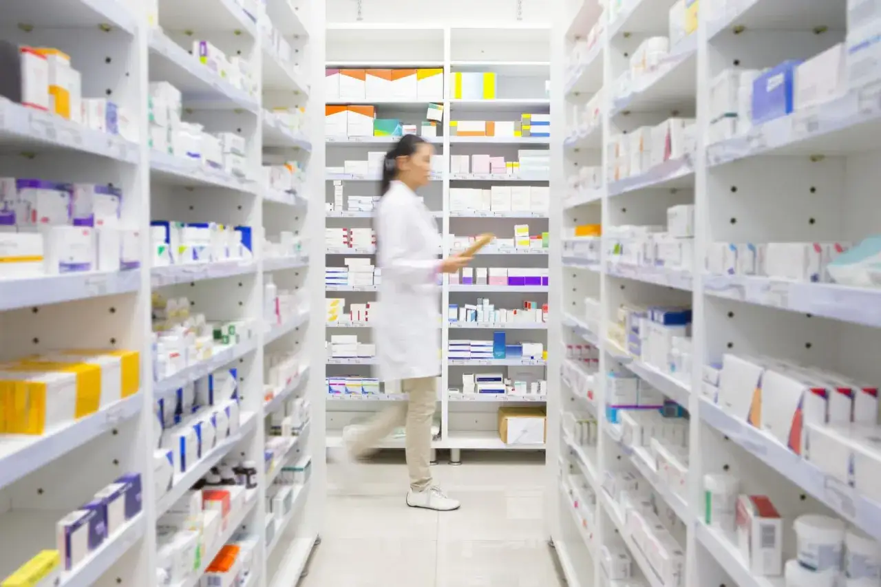 Farmaceutka w białej kitli przechodzi między regałami pełnymi leków. Pytanie 