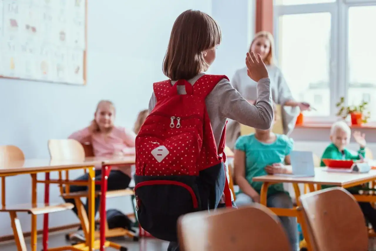 Dziewczynka z plecakiem wita się z nauczycielką. Wskazówki, jak postępować z dzieckiem z fobią szkolną, mogą pomóc w takich sytuacjach.